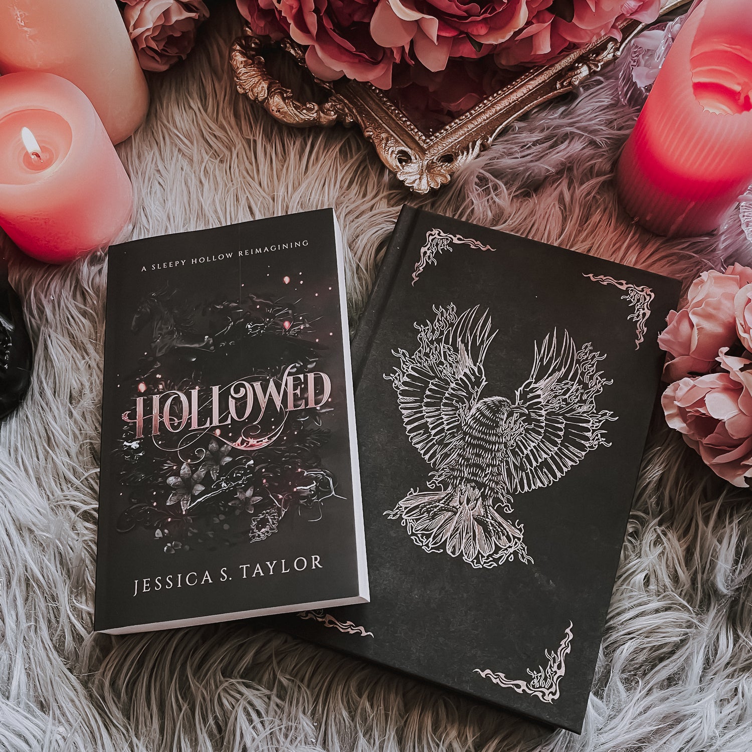 Image of a paperback resting at an angle atop a hardback. The paperback is titled HOLLOWED and features a collection of items behind the swirling fire styled typography. The hardback is a black hardback with an illustrated firebird in the center and decorative corners stylized in flames. The background is a gray fur rug, and pink flowers and candles surround the border.