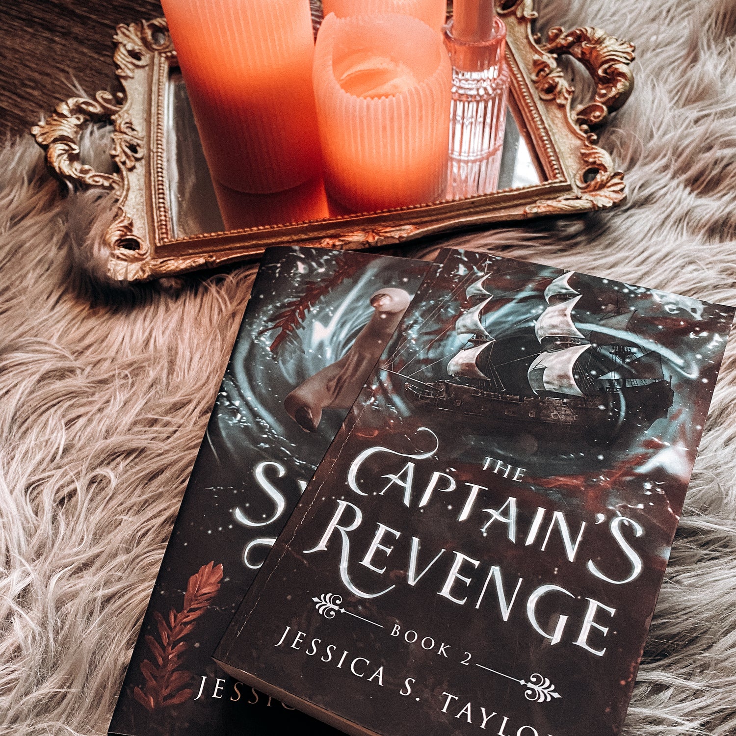A paperback of The Captain's Revenge, featuring a ship in a teal ocean with red blood spreading through the water, sits atop a hardback of The Syren's Mutiny. In the background, a gray fur rug. In the top border area, a mirrored tray with pink candles.